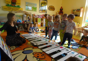 Dzieci grają w zespole na Kolorpiano, stawiają stopę na dany klawisz zgodnie z instrukcją nauczyciela, który wskazuje dany dźwięk na tablicy.